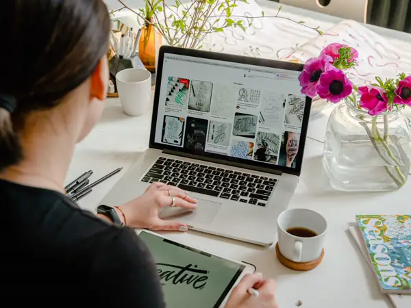 A person working at a laptop on a bright desk with flowers and a coffee